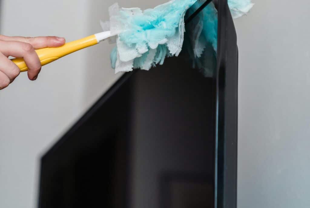Close-up of a hand using a duster to clean dust from the top edge of a monitor in an office setting.