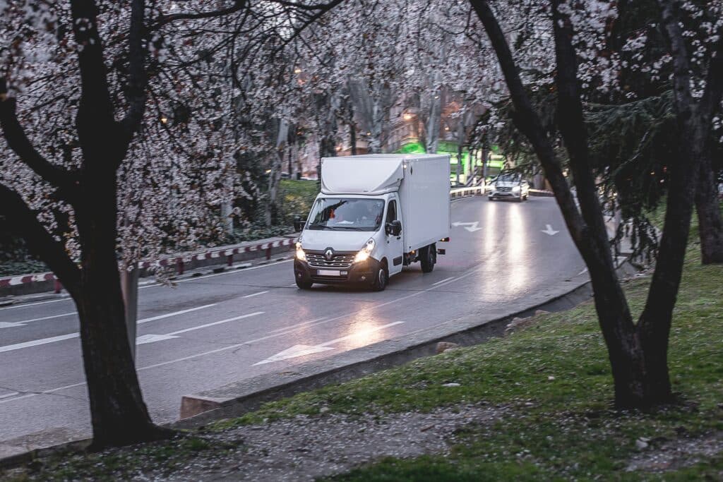 White delivery truck driving on a city road surrounded by trees, illustrating travel for a cleaning business.