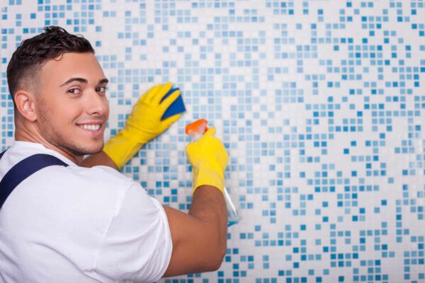 Smiling young male janitor cleaning blue mosaic tiled wall with a spray bottle and yellow gloves.