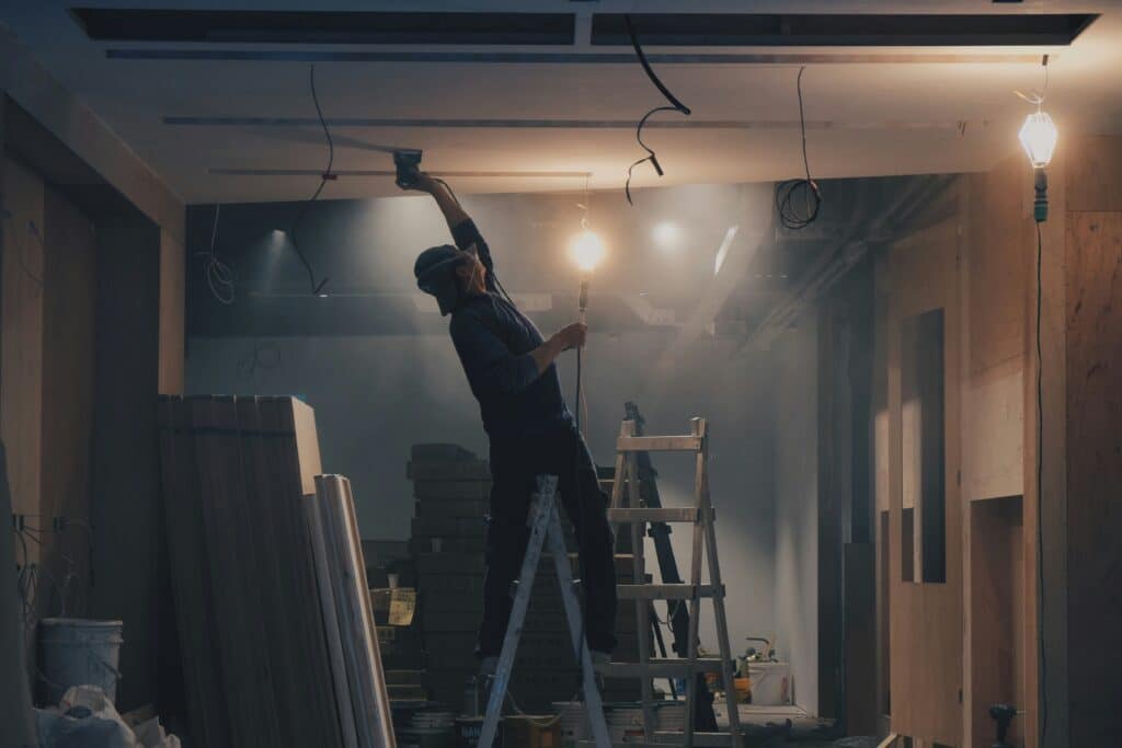 A worker standing on a ladder sanding a ceiling in a construction site with dim lighting, surrounded by construction materials and debris.