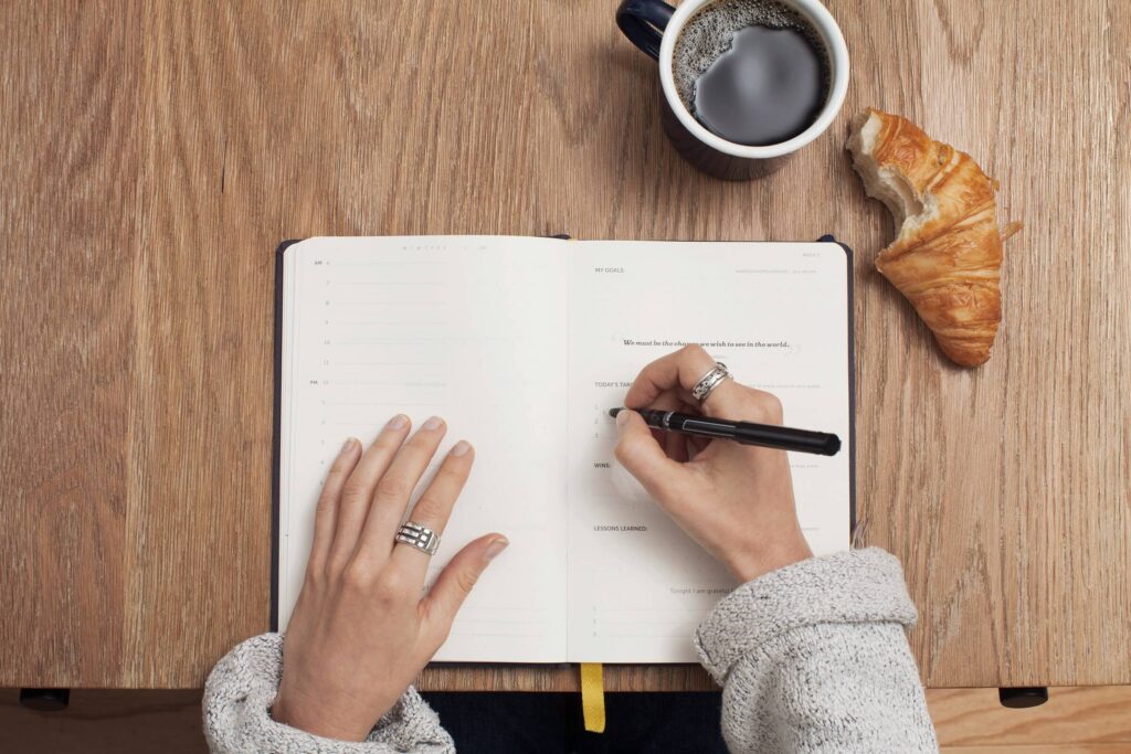 A person writing in a notebook next to a coffee cup and croissant, symbolizing organized planning and productivity.