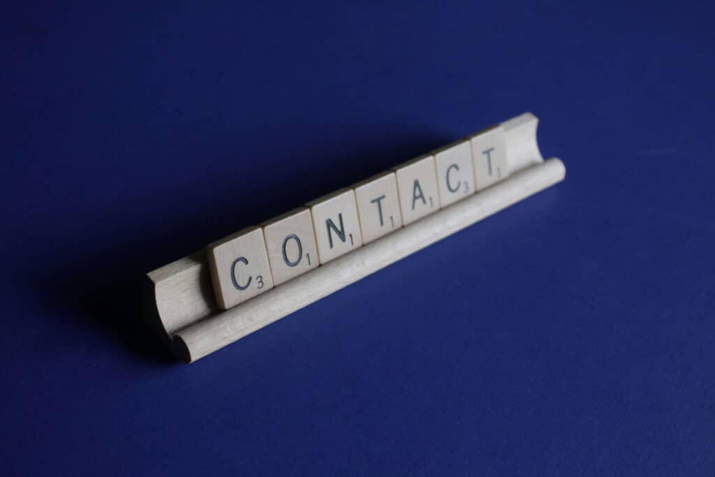 Scrabble tiles spelling "Contact" on a wooden tile holder, set against a blue background.