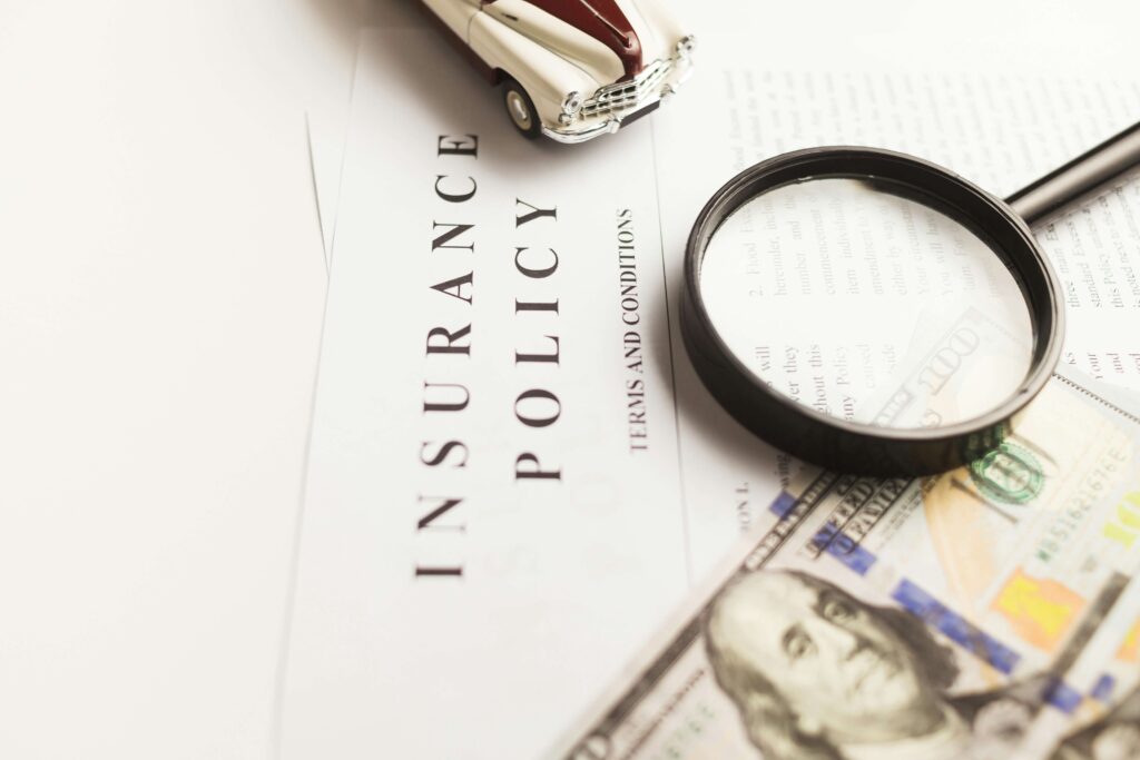 A magnifying glass over an insurance policy document with a hundred-dollar bill and a vintage car model in the background.